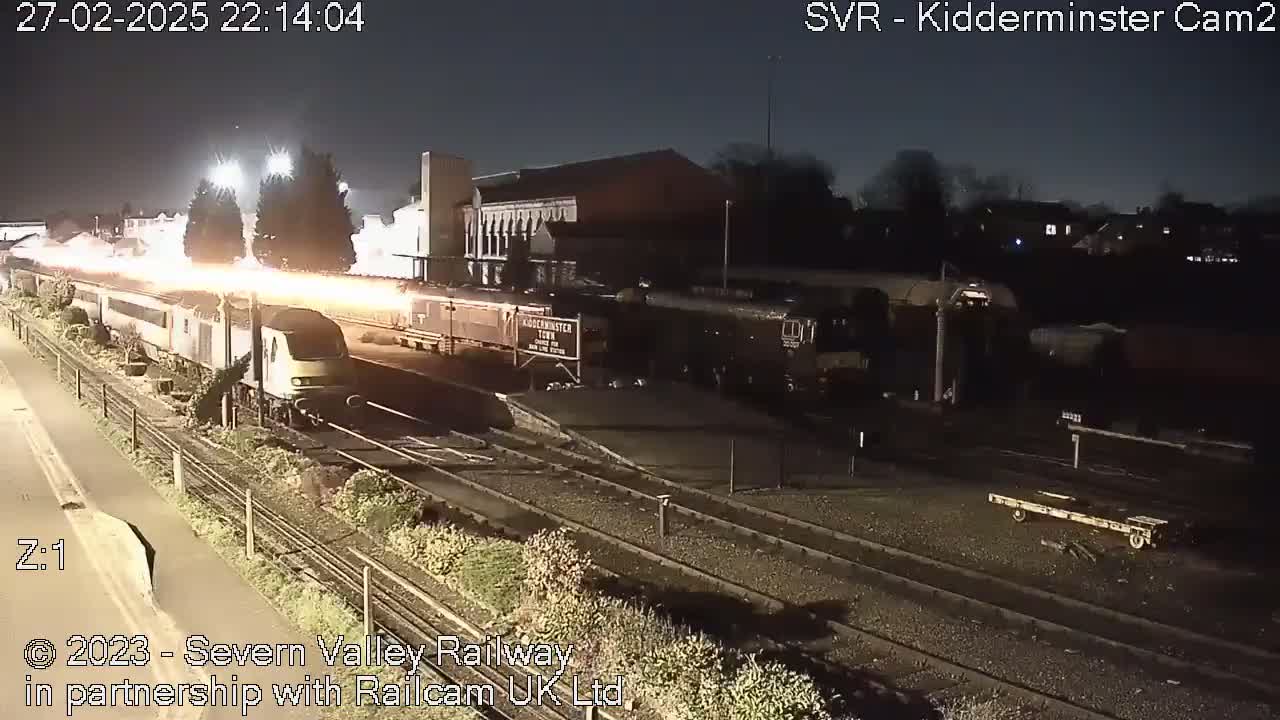 At night, a train departs a station, its headlights illuminating the tracks and surrounding area.