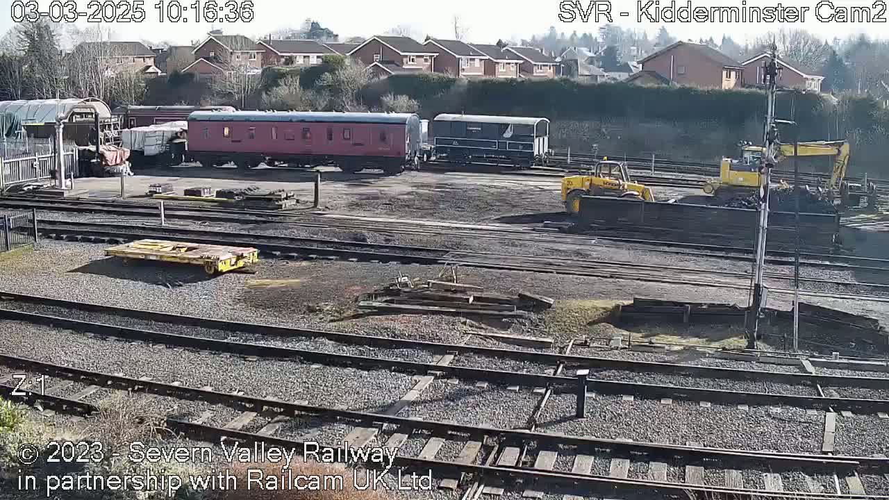 A sunny day at a railway yard shows several rail cars, two yellow construction vehicles, and multiple sets of train tracks.