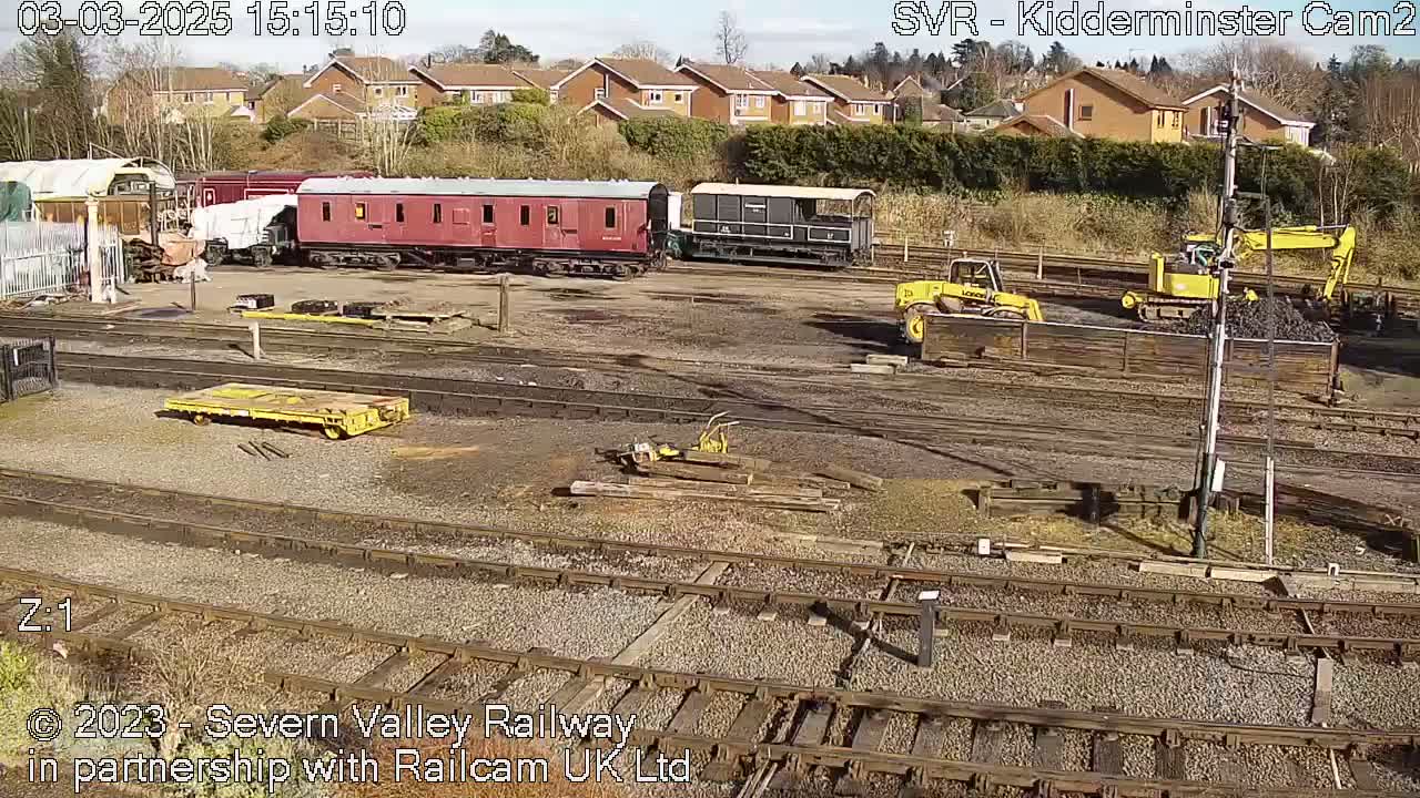 A sunny day at a railway yard shows several train cars, yellow construction equipment, and railway tracks.