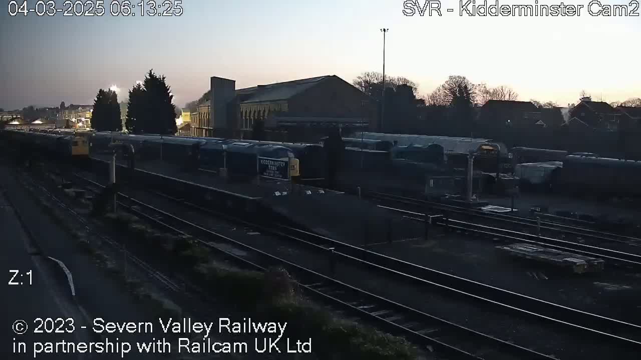 Under a dusky pre-dawn sky, numerous train cars are parked on a railway track beside a building.