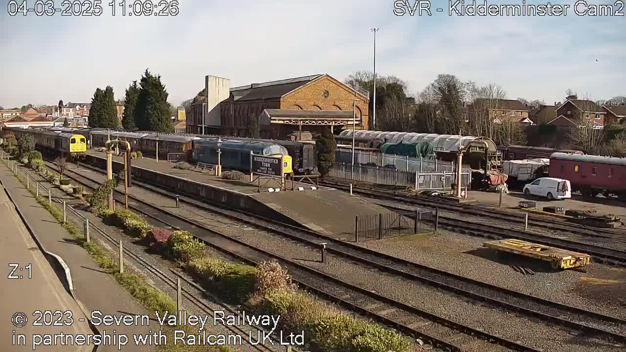 A sunny day at a railway station shows numerous train cars and locomotives parked on multiple tracks, with a brick building in the background.