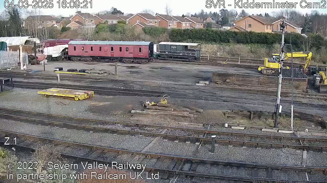 A railway yard with several railcars, a yellow excavator, and a pile of coal is visible under an overcast sky.