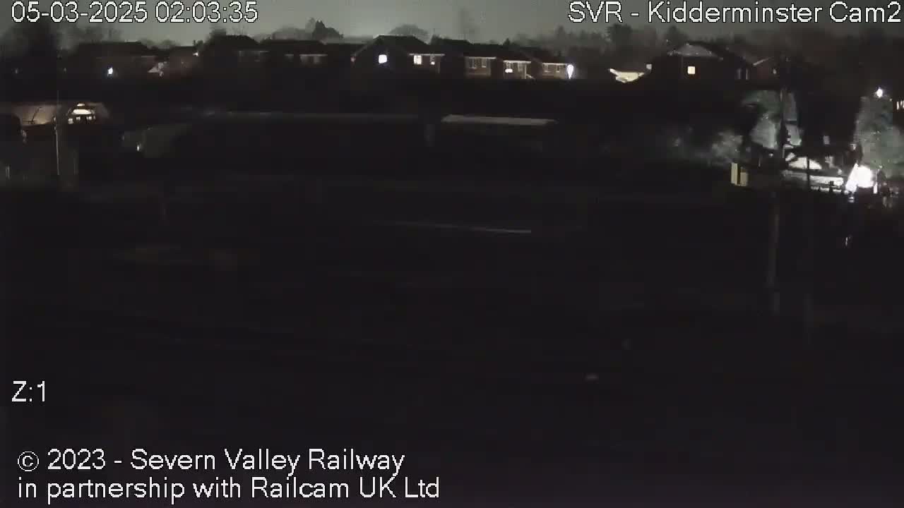 A nighttime, dark view of a railway line with some buildings in the distance under a cloudy sky.