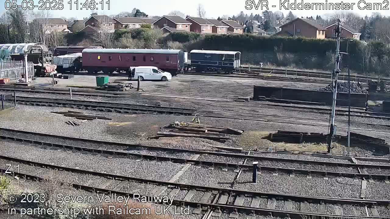 A sunny day at a railway yard shows several railway carriages and a van parked near railway tracks, with residential buildings visible in the background.