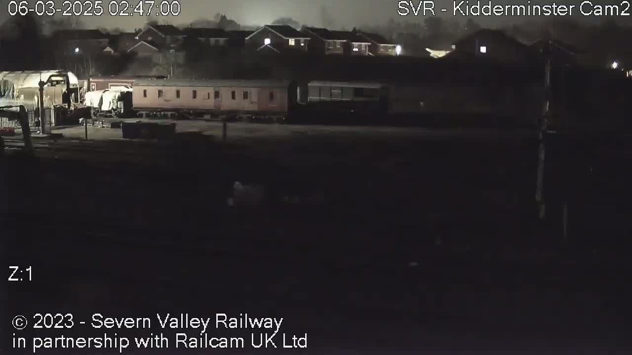 A dimly lit nighttime scene shows a train and railway cars parked near residential buildings under a cloudy sky.