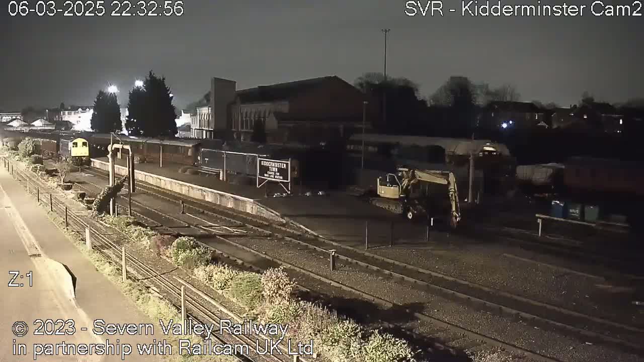 A nighttime view of a train station with a train parked on the tracks, a small excavator nearby, and a building in the background.