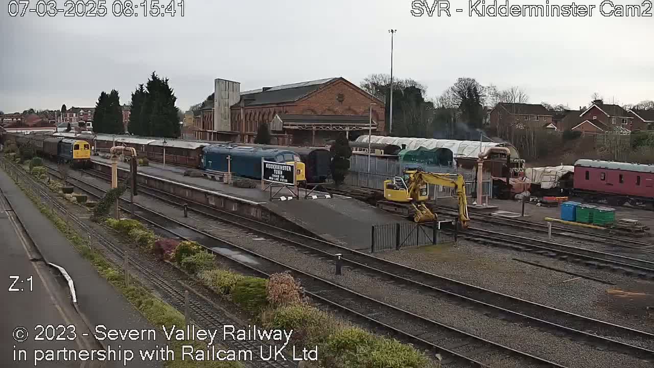 A train station with numerous train cars and a small excavator on an overcast day.