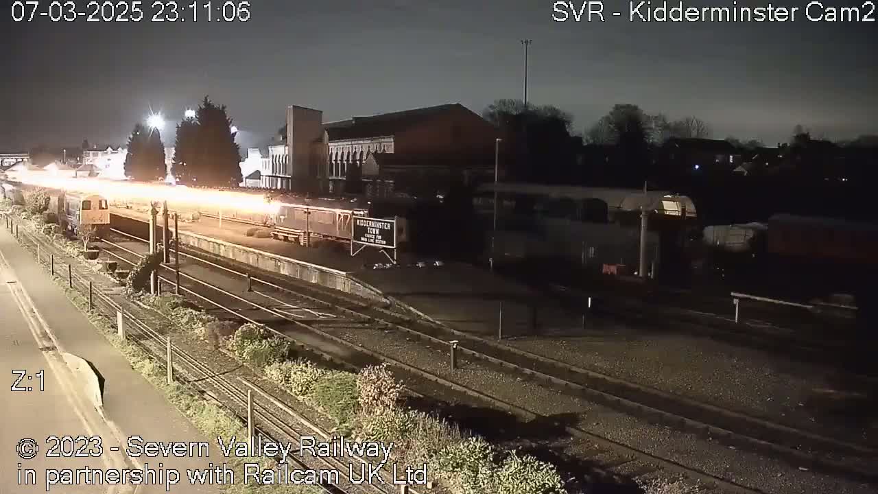 At night, a train with bright lights passes through a dimly lit train station.