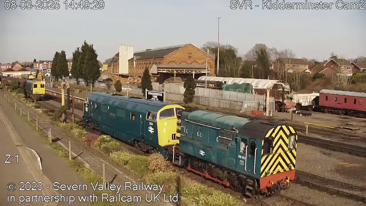 Two teal and yellow diesel locomotives are parked at a train station on a sunny day, with other train cars and buildings visible in the background.