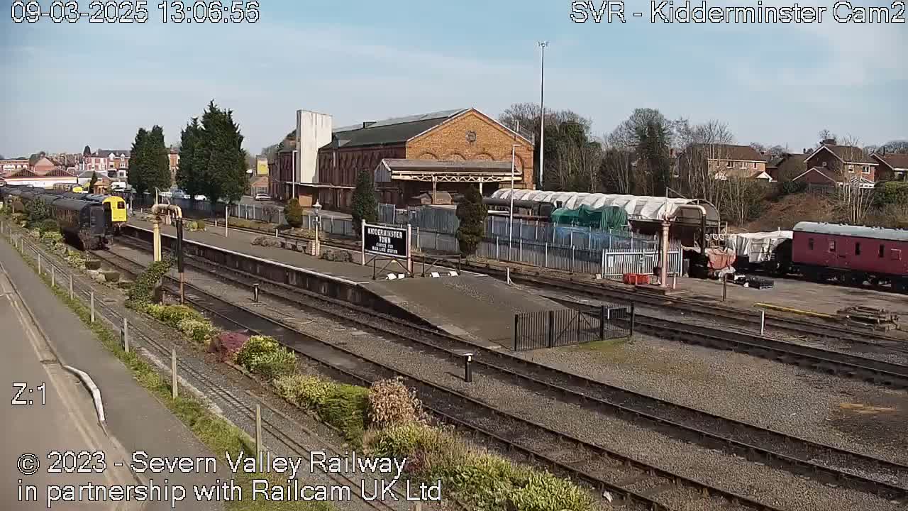 A sunny day at a train station shows several rail cars, a brick building, and railway tracks.