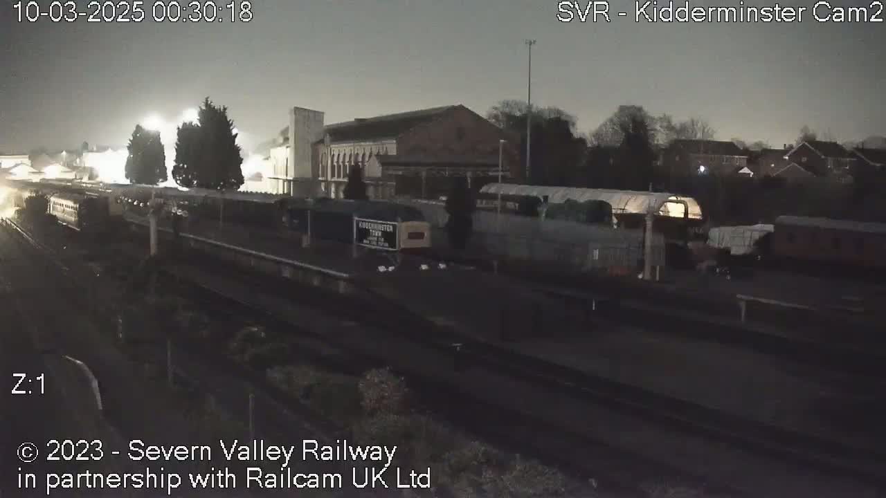 A nighttime view of a train station with several railway cars and buildings under a dark, possibly overcast sky.