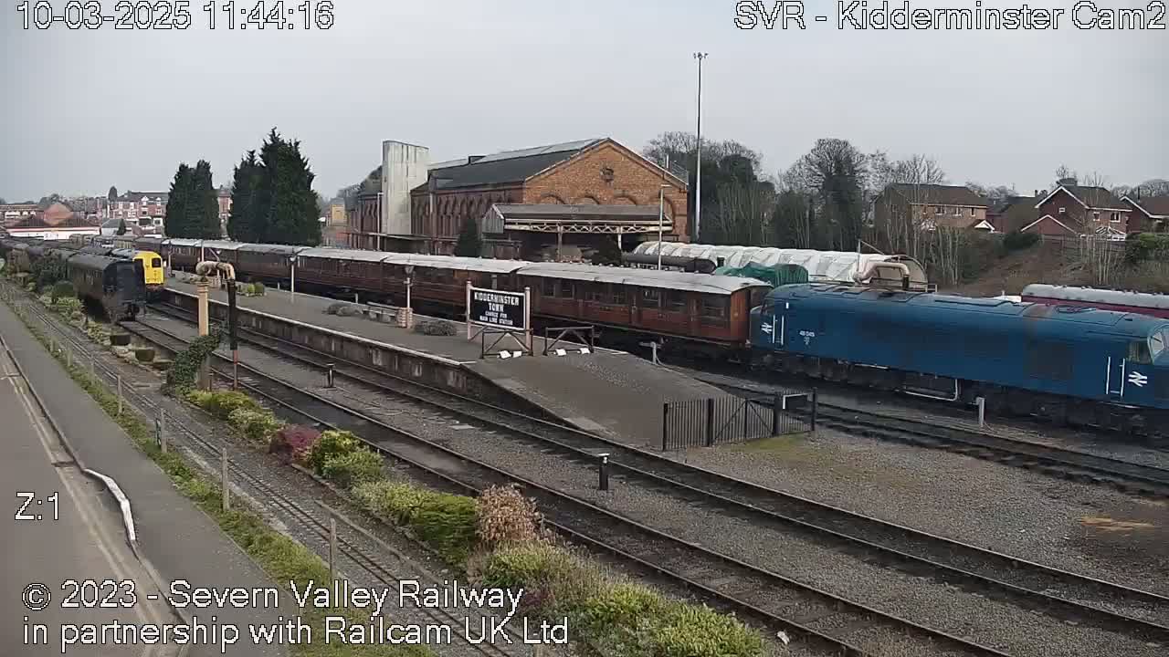 A long passenger train is at a station, with a blue diesel locomotive at the end of the track under an overcast sky.