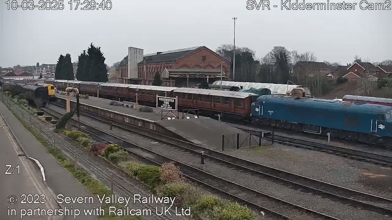 A train station on an overcast day with several railway carriages and a blue diesel locomotive on the tracks.
