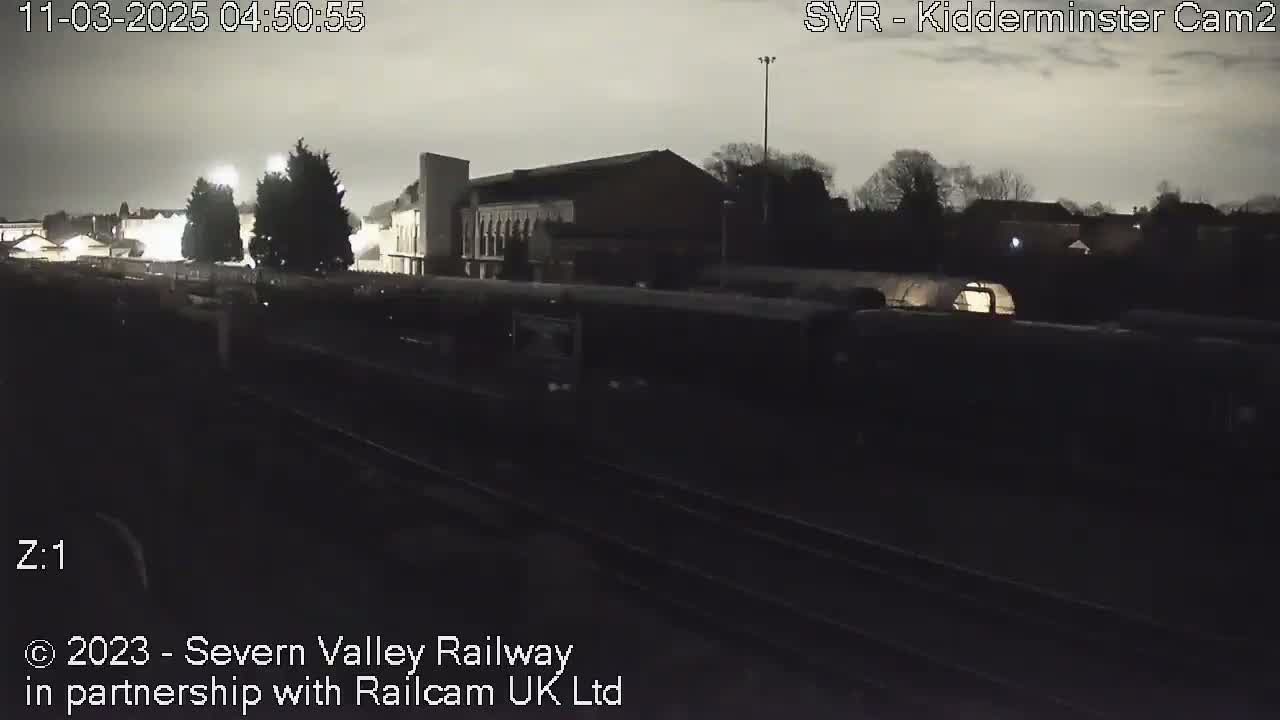 A dark, nighttime view of a train moving past a station building under a cloudy sky.