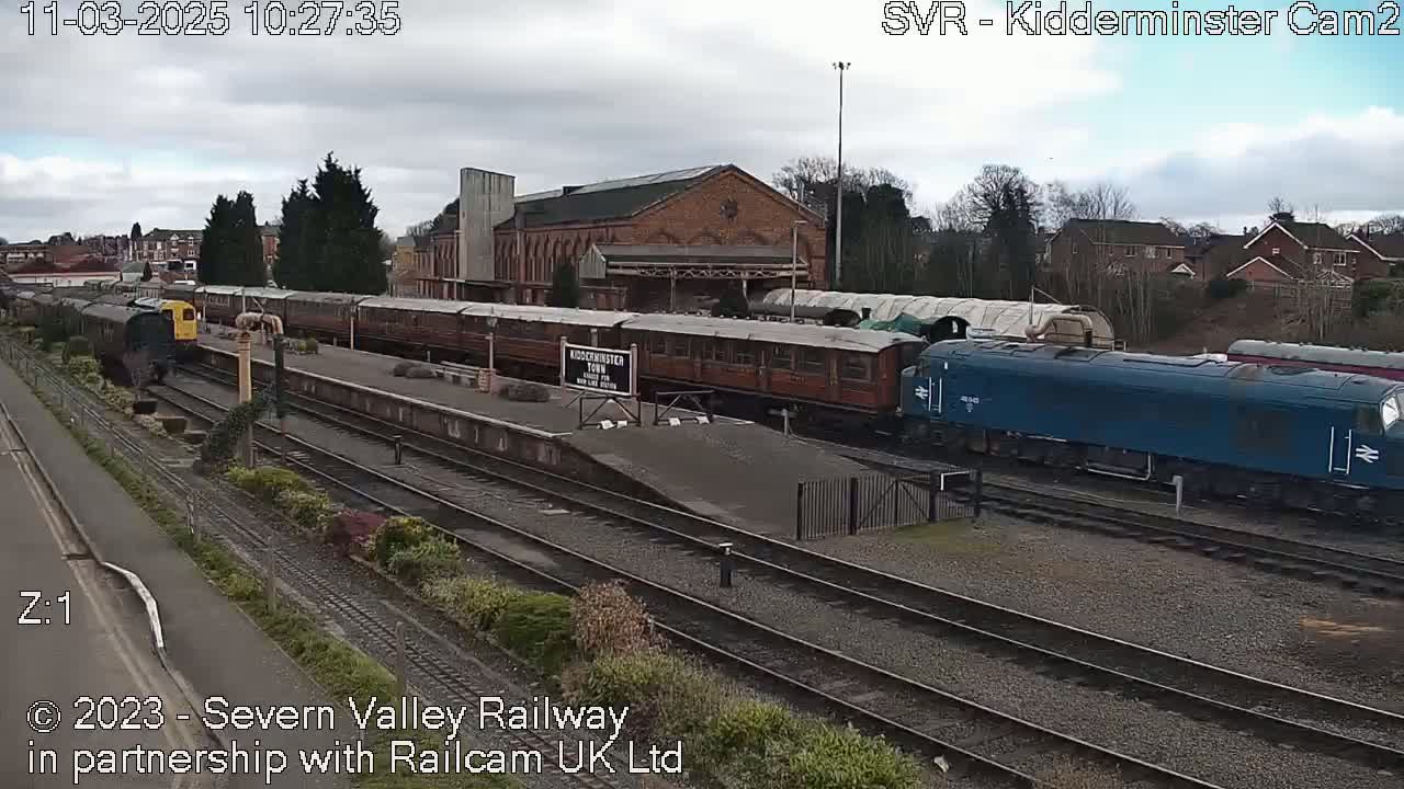 A blue diesel locomotive and a long train of brown passenger cars sit in a train station on an overcast day.