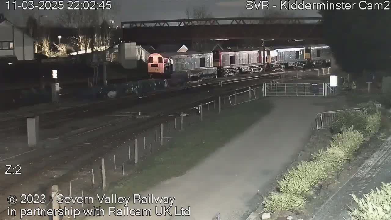 At night, a train consisting of several locomotives and cars sits under a bridge near some shrubs and a fence.