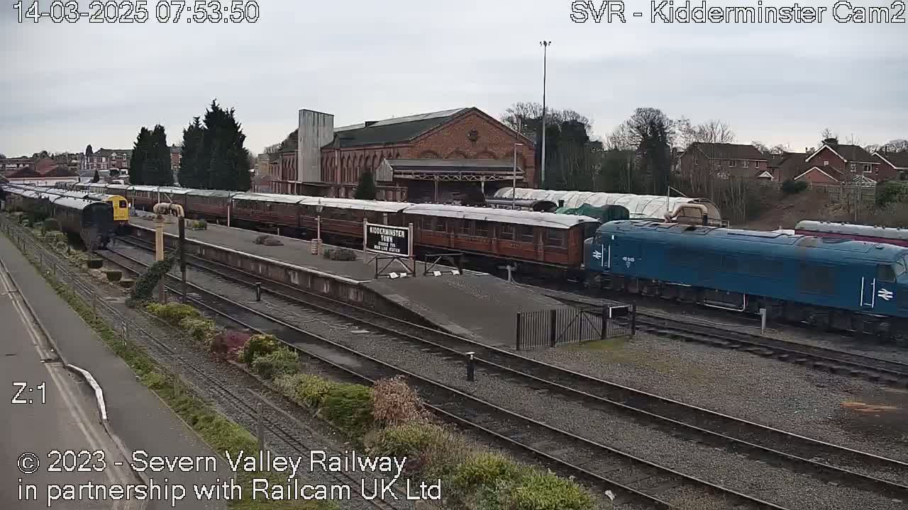 A train station on an overcast day with multiple passenger cars and a blue diesel locomotive visible on the tracks.