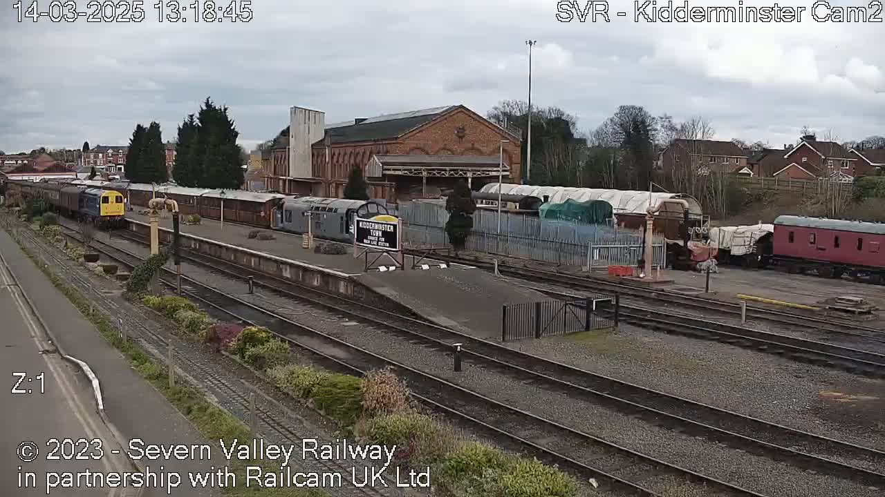 A train station with several railway cars and a brick building under an overcast sky.
