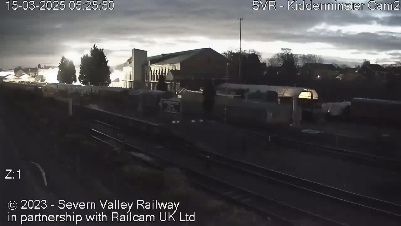 A train passes by a station under an overcast sky at dawn.