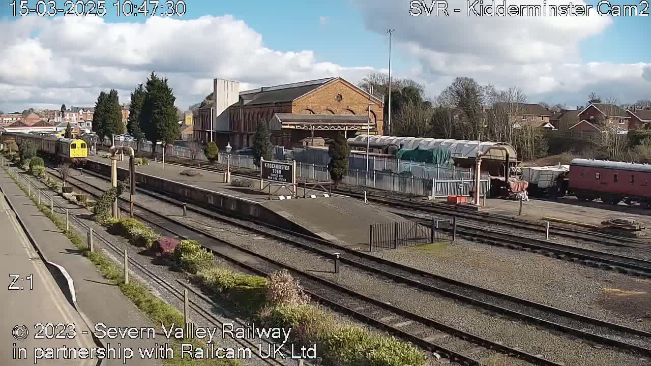 A mostly sunny day at a train station shows a yellow train on the tracks and several other train cars parked nearby.
