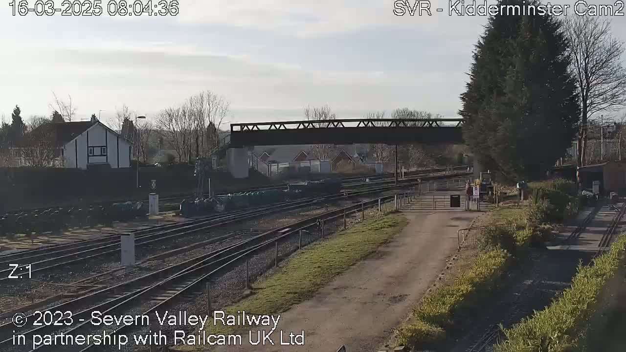 A railway yard with several train tracks, a bridge overhead, and some buildings in the background under a mostly clear sky.