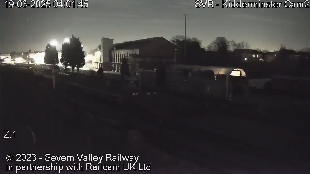 A nighttime view of a train station area under a dark sky, with some buildings and trees visible.