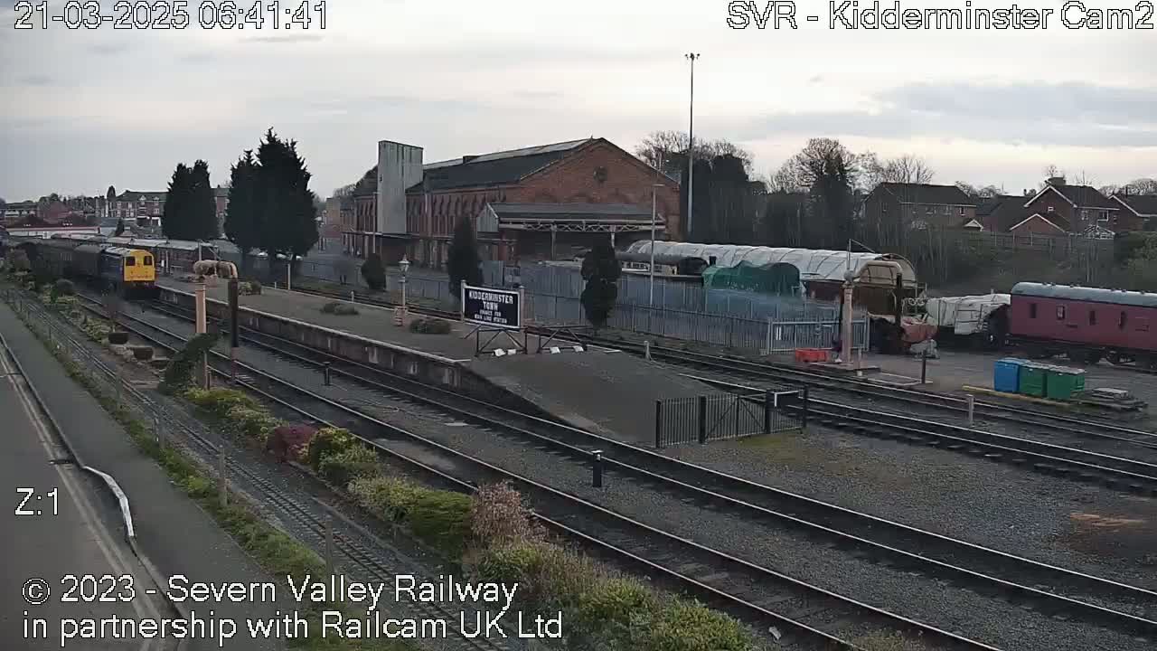 A diesel locomotive leads a train past a station platform under an overcast sky.