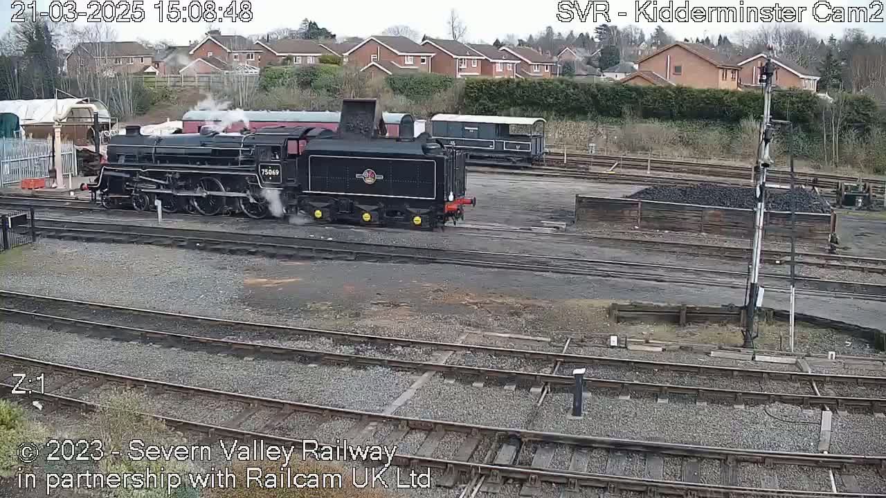 A black steam locomotive with plumes of smoke is parked on train tracks in a rail yard next to other rail cars on an overcast day.