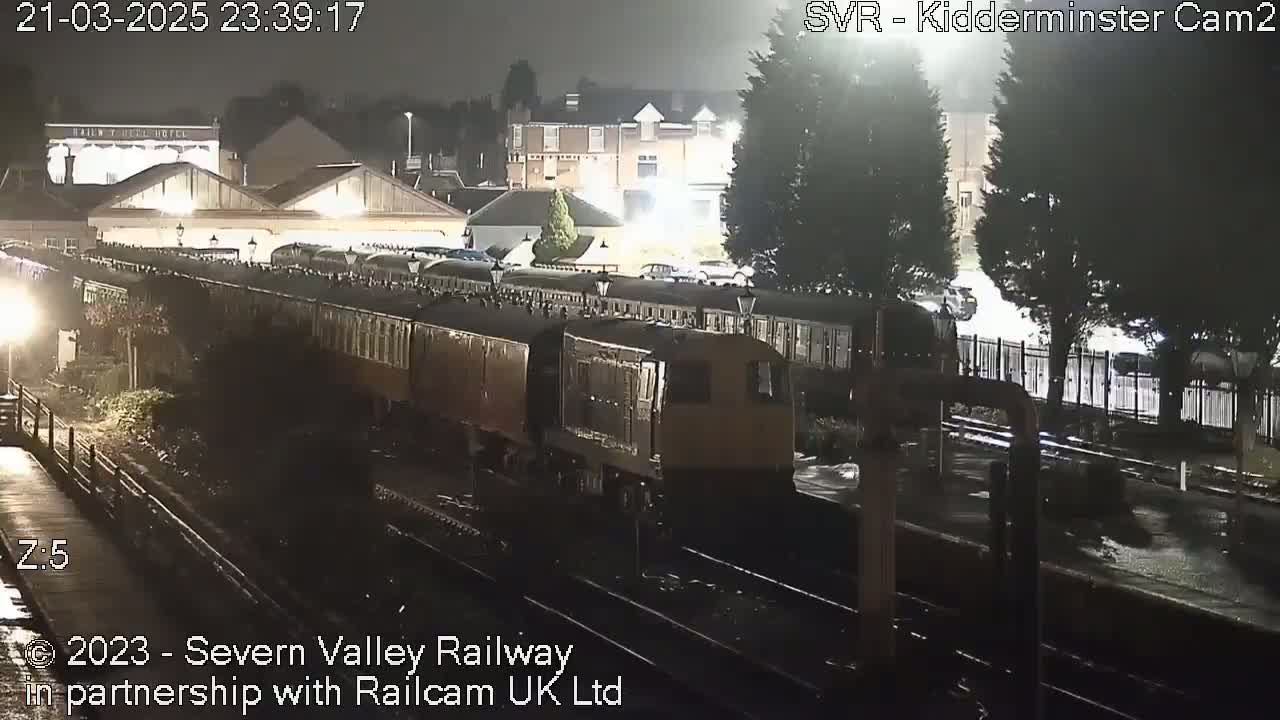 At night, under wet conditions, a long brown train sits in a station next to some buildings and trees.