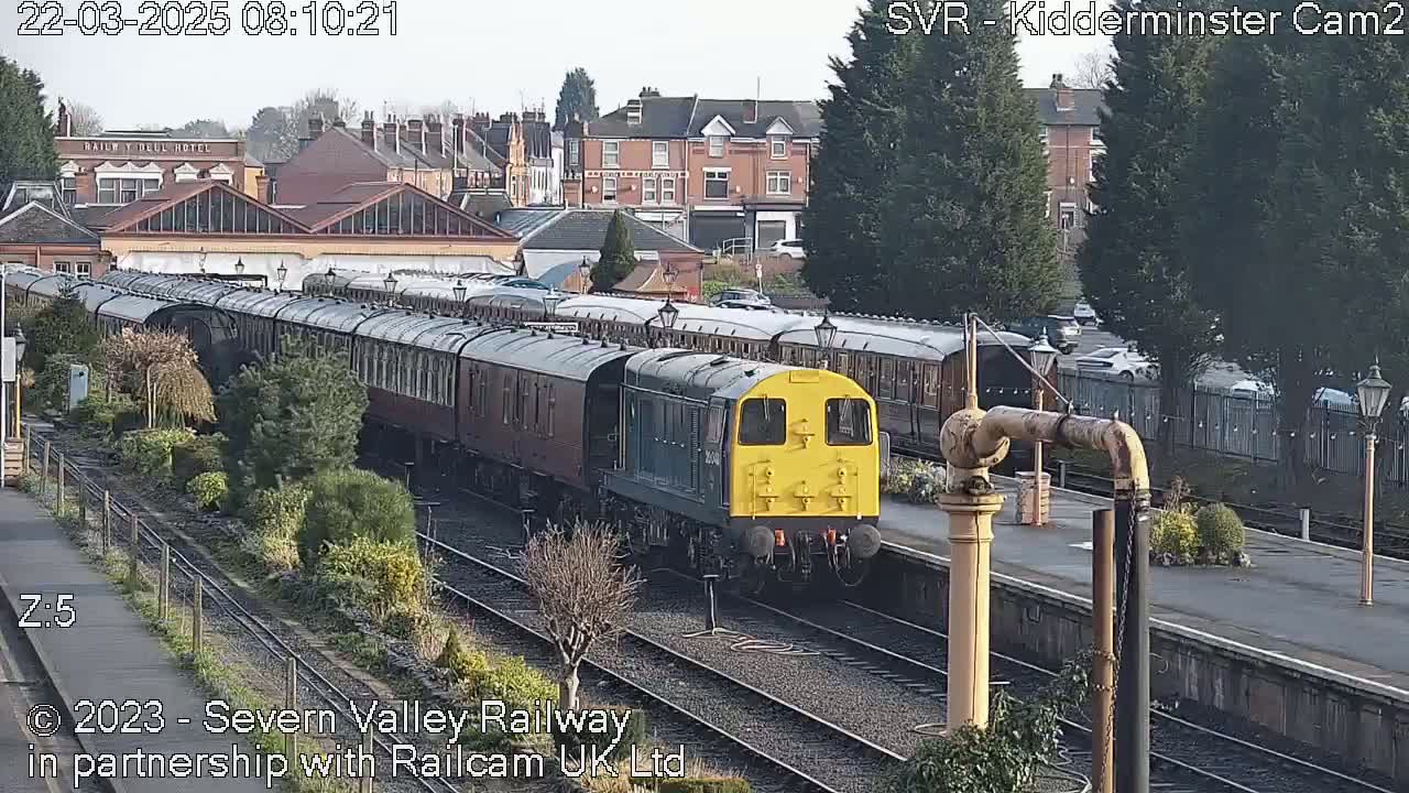 A diesel locomotive pulls a long train of passenger cars through a railway station on a sunny day.