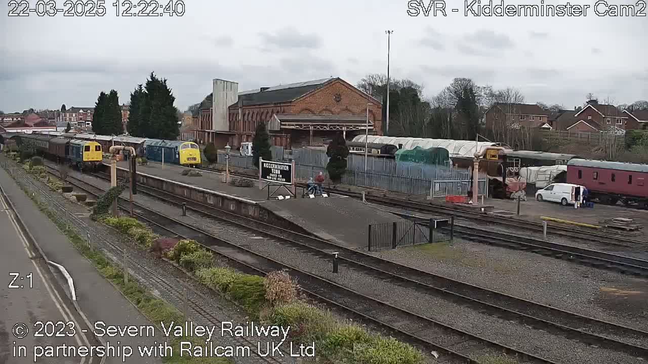 A train station with multiple train cars and a brick building is visible on an overcast day.