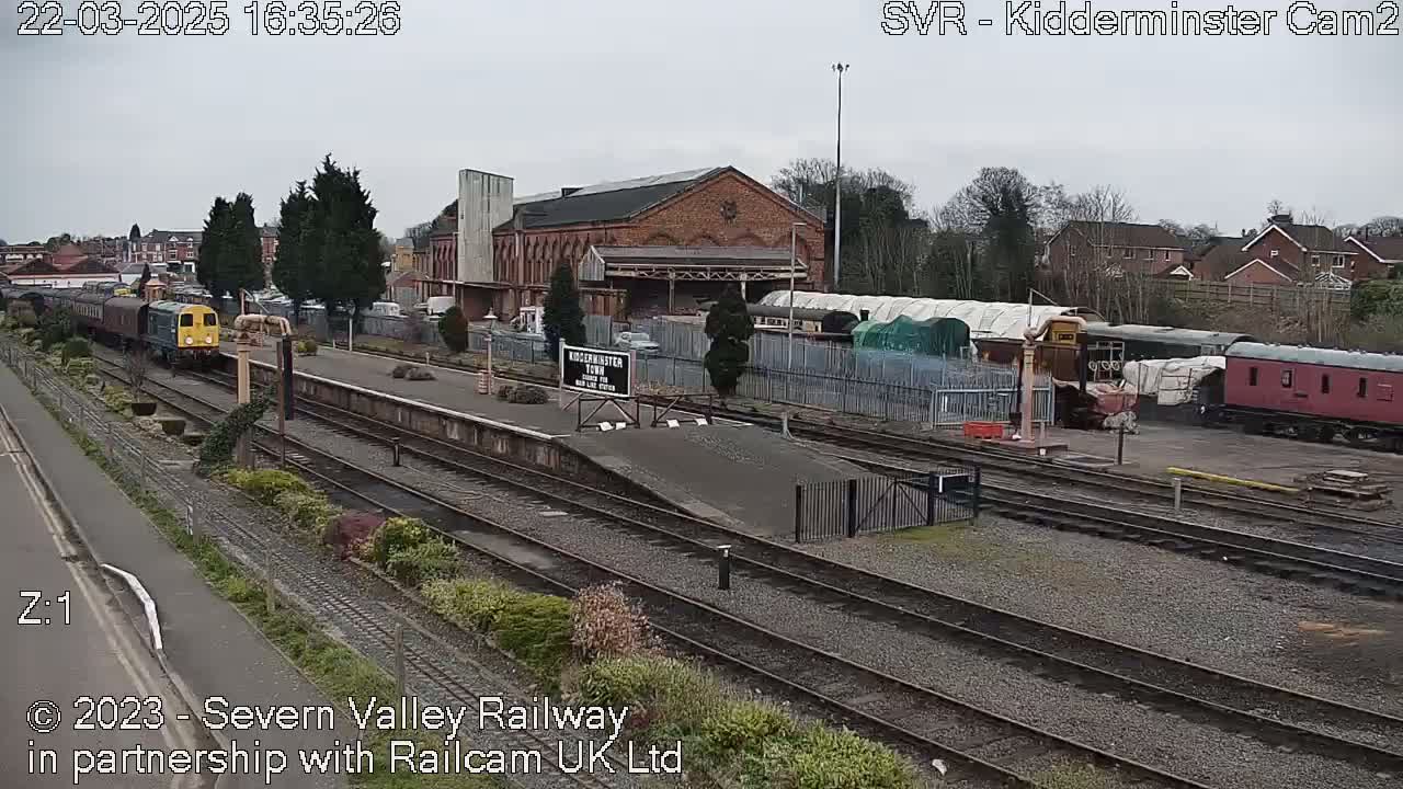 A train pulls into a railway station on an overcast day, with several other rail cars visible in the station yard.