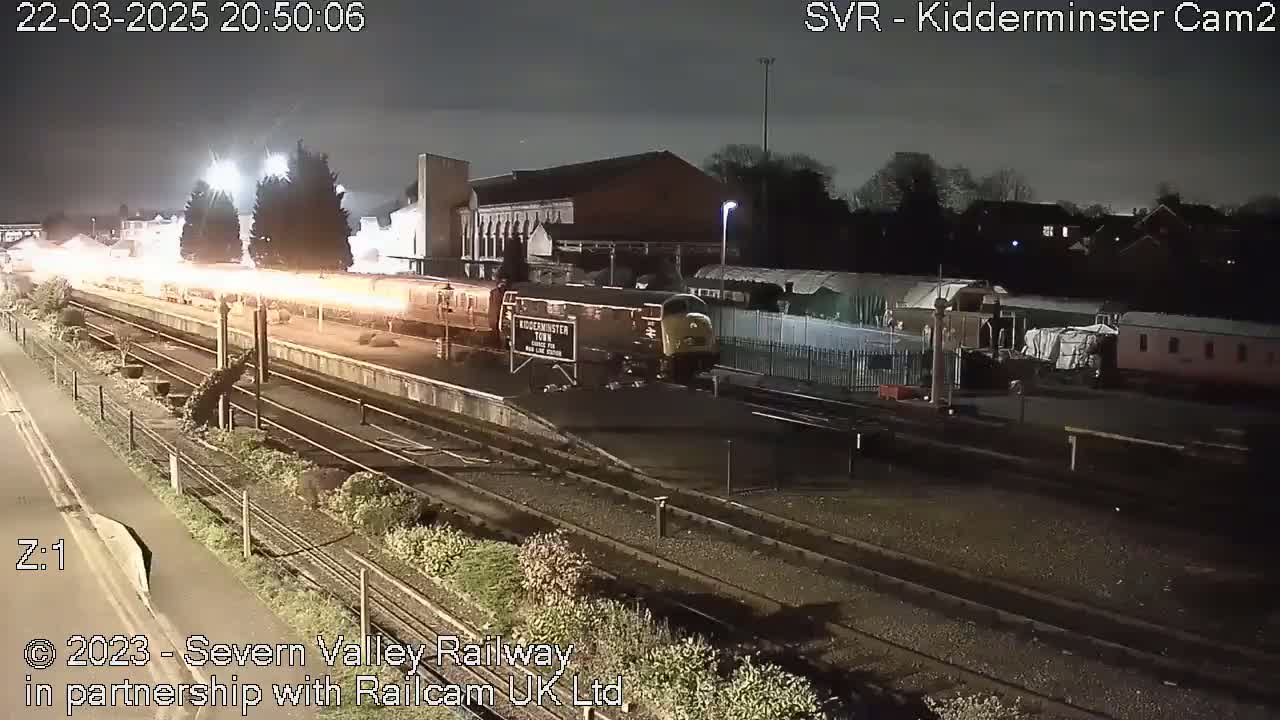 A nighttime view of a train station with a diesel locomotive and several railway cars parked on the tracks under dark, cloudy skies.
