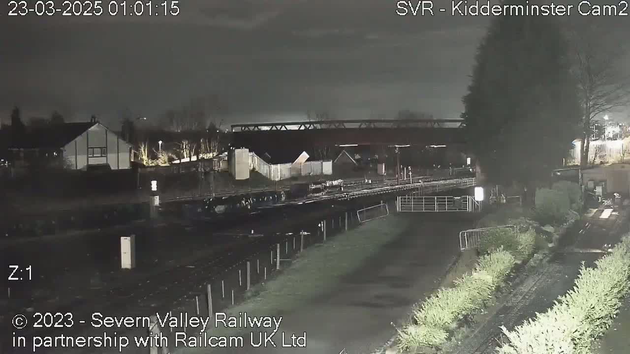 A nighttime view of a canal and railway line under a dark sky, with houses and trees visible in the distance.