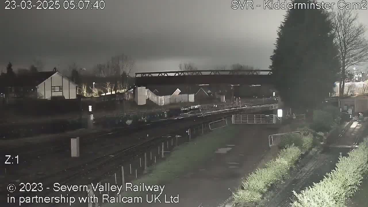 A nighttime view of a railway line, passing under a bridge and alongside houses, in overcast conditions.