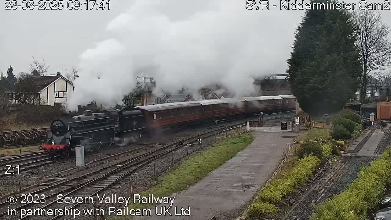 A steam train with multiple passenger cars chugs along train tracks on an overcast day, emitting a large plume of white smoke.