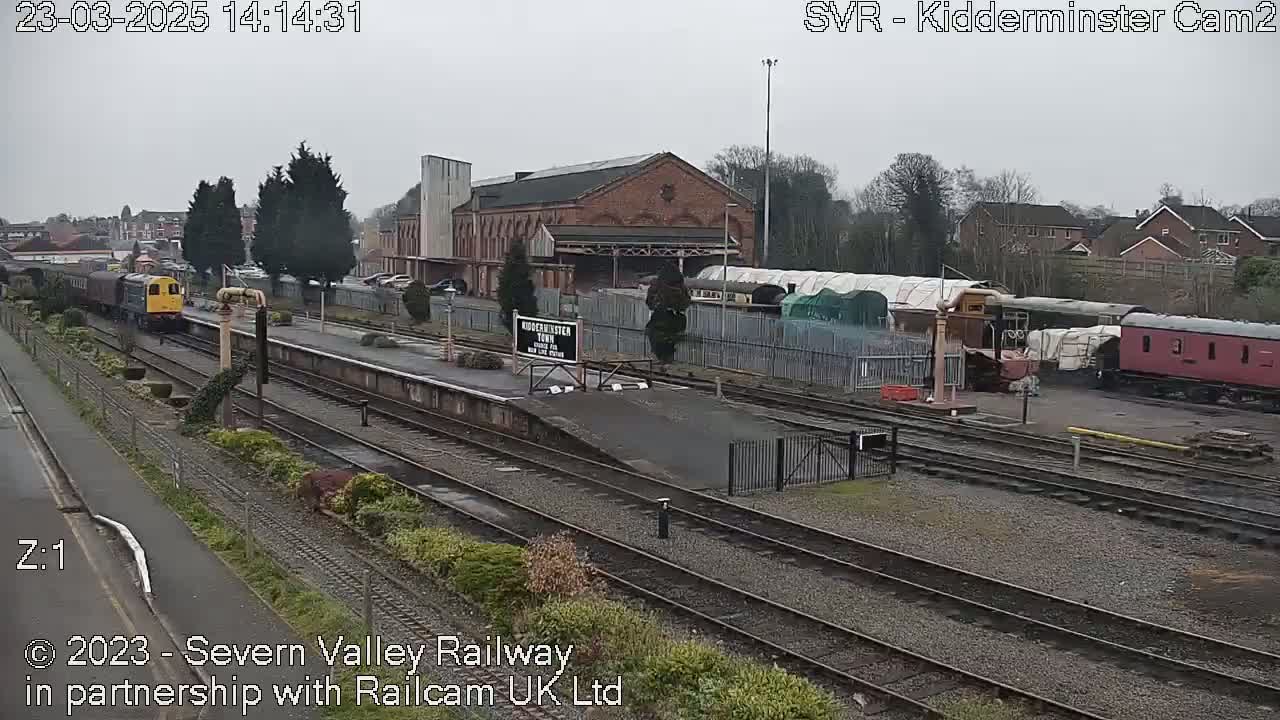 A diesel train pulls away from a station platform on an overcast day, passing several rail cars and a brick building.