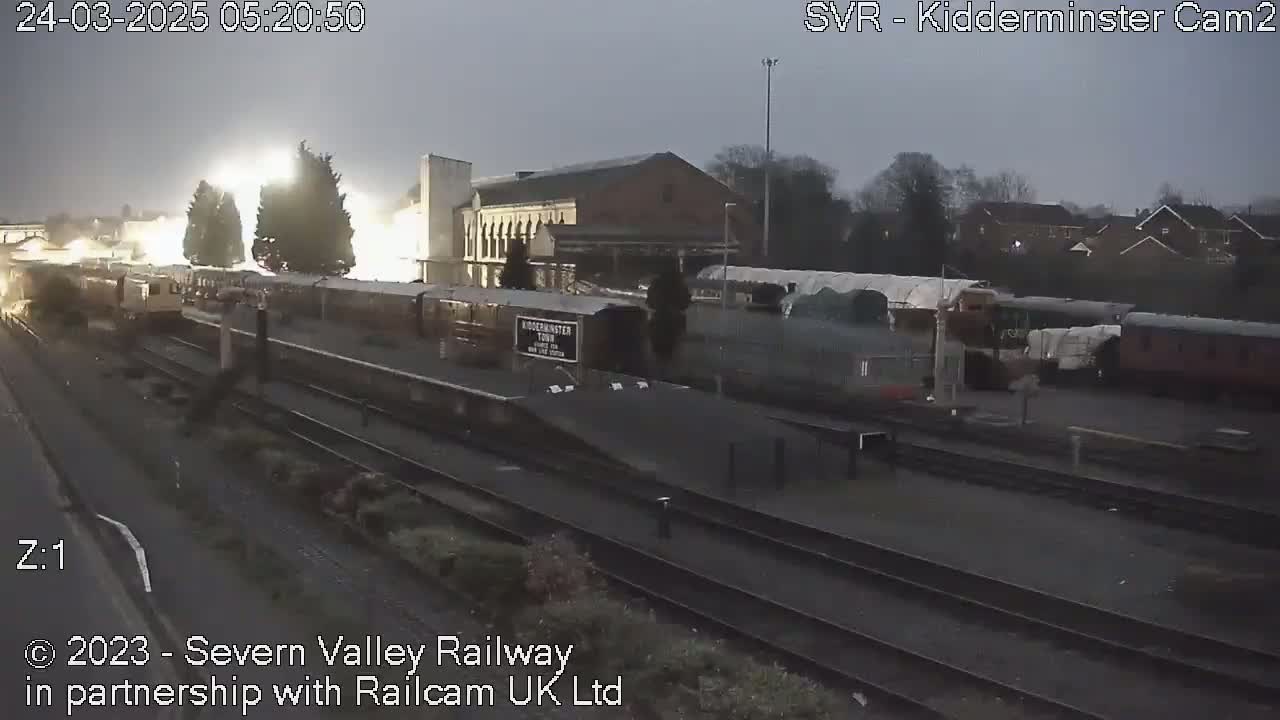 A train station at twilight, with several trains parked on the tracks under bright artificial lights.