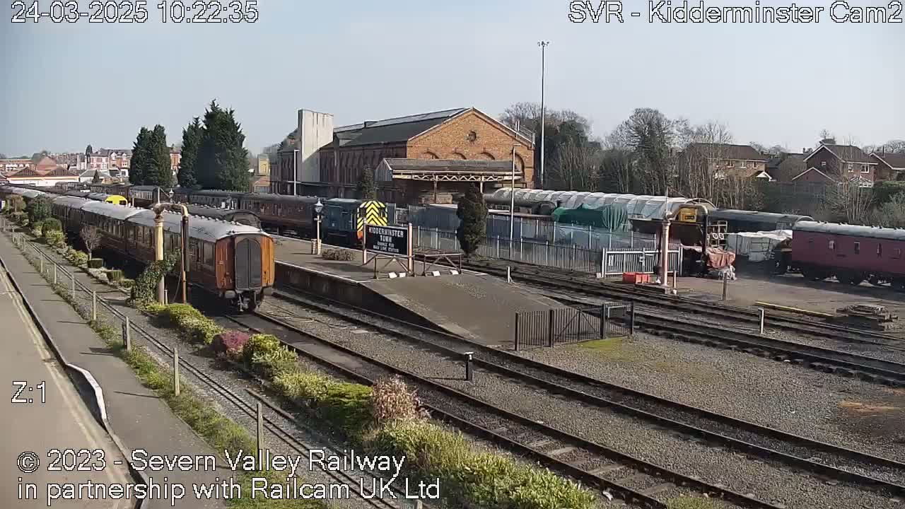 A sunny day at a train station shows several passenger cars and locomotives on multiple tracks, with a brick building in the background.