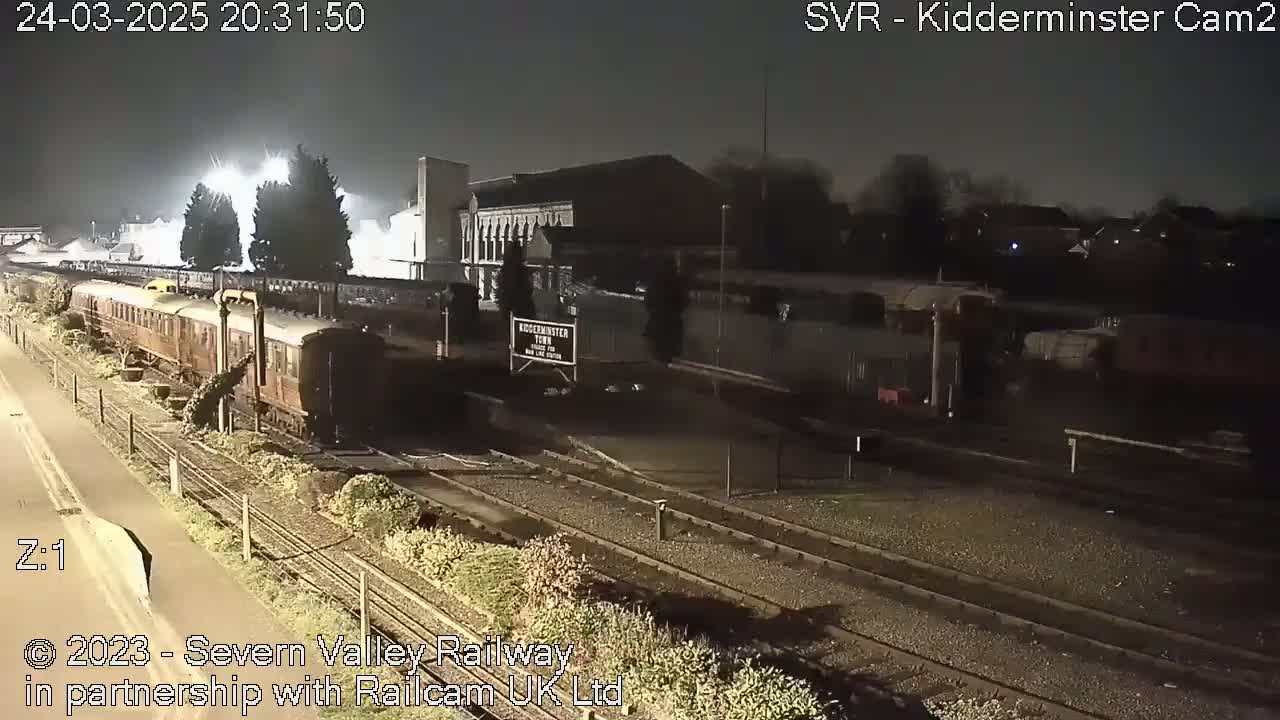 A nighttime view of a train station with a passenger train on the tracks under artificial light.