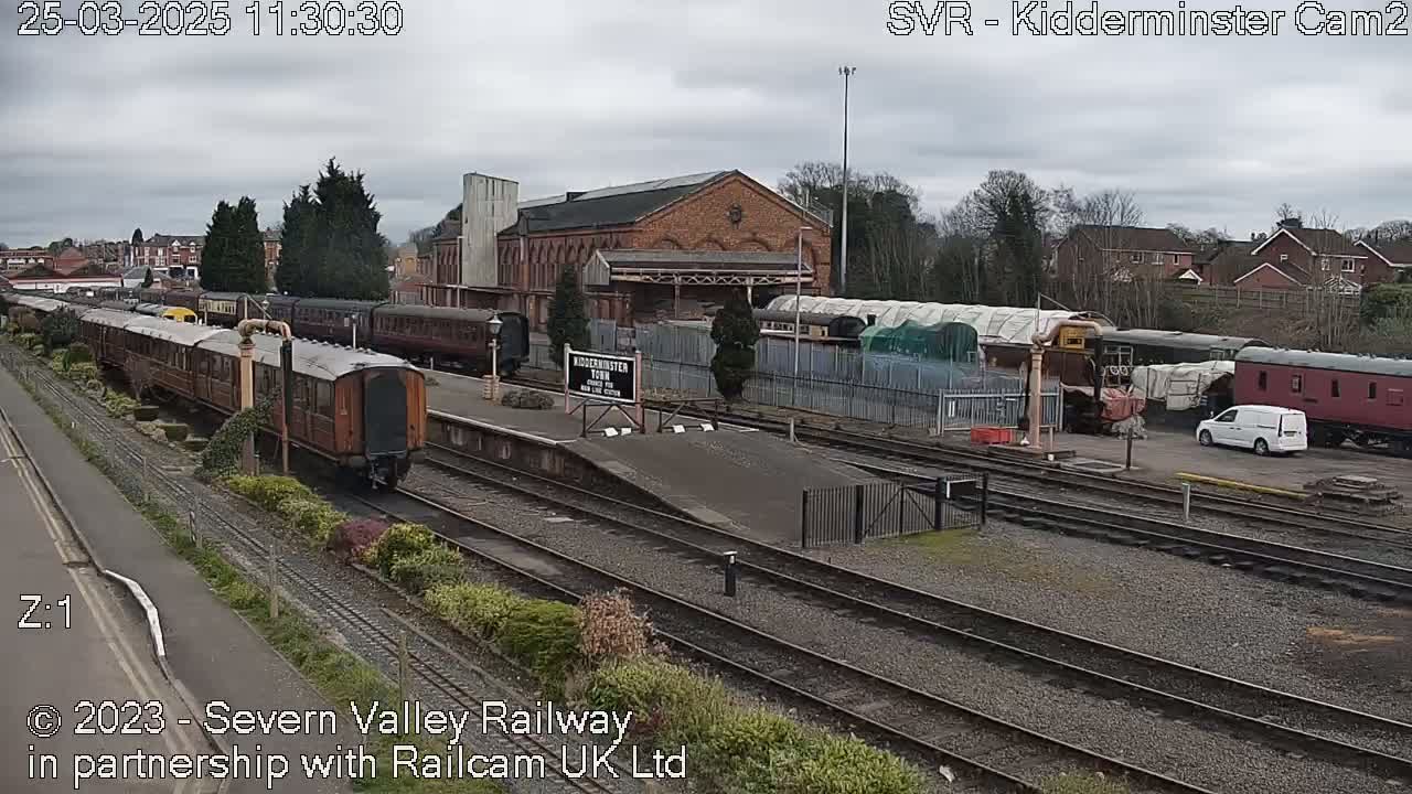 A train station with several train cars and a brick building is visible under an overcast sky.
