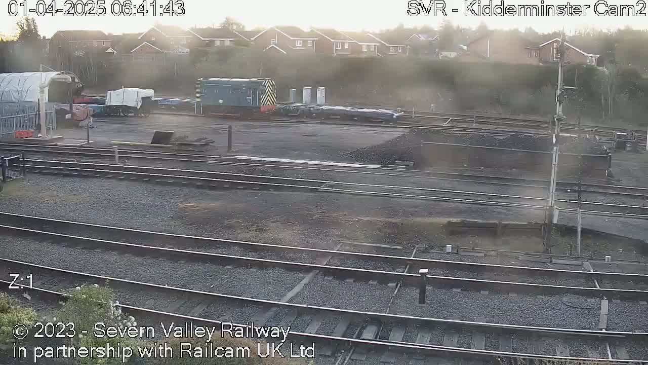 A hazy sunrise illuminates a railway yard with several train cars and locomotives, including one dark-green diesel locomotive, near a residential area.