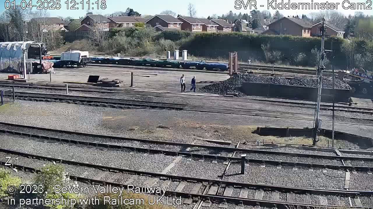 A sunny day at a railway yard shows several railcars, a pile of coal, and two people walking near the tracks.