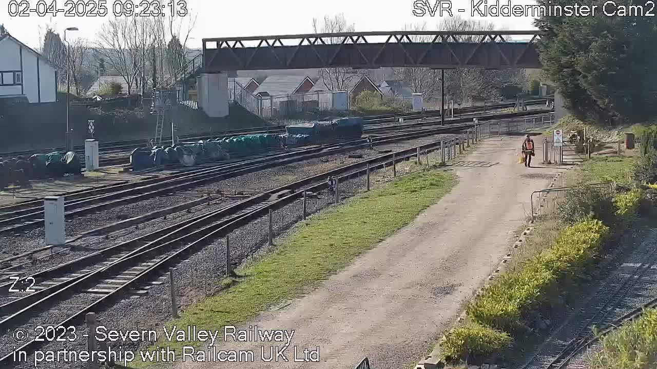 A person walks along a dirt path next to a railway yard on a sunny day.