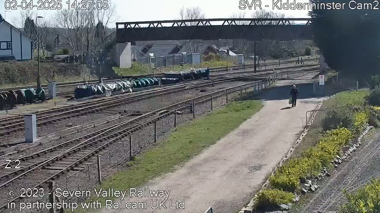 A person walks along a gravel path next to railroad tracks on a sunny day, with several covered railcars visible in the background under a bridge.