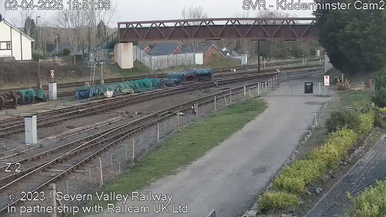 A gravel path leads to a railway yard with several covered train cars parked on multiple tracks under a metal bridge, all under an overcast sky.