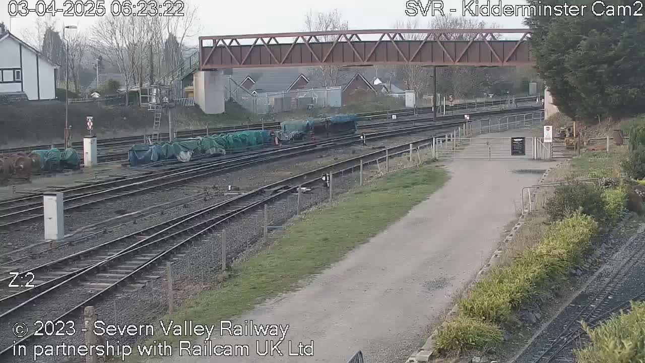 A railway yard with several train cars covered in green tarps sits under a metal bridge on an overcast day.