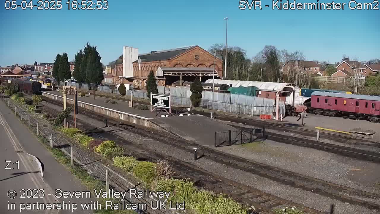 A sunny day at a railway station shows several train cars, a brick building, and train tracks.