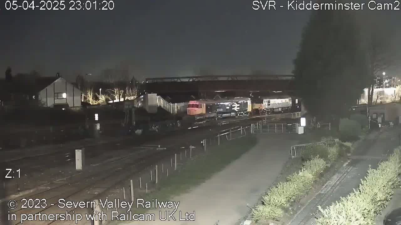 Several diesel locomotives are parked at night next to a railway line and a paved path, under a dark sky.