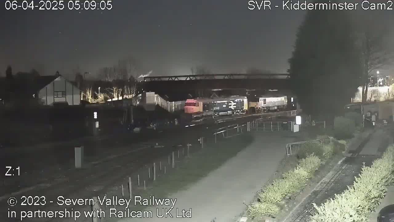 Several locomotives are parked at night near a railway line under a bridge, next to some vegetation and residential buildings.